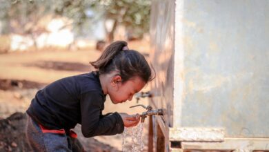 A little girl drinks water from a faucet outdoors in Idlib, Syria, representing hygiene and innocence.